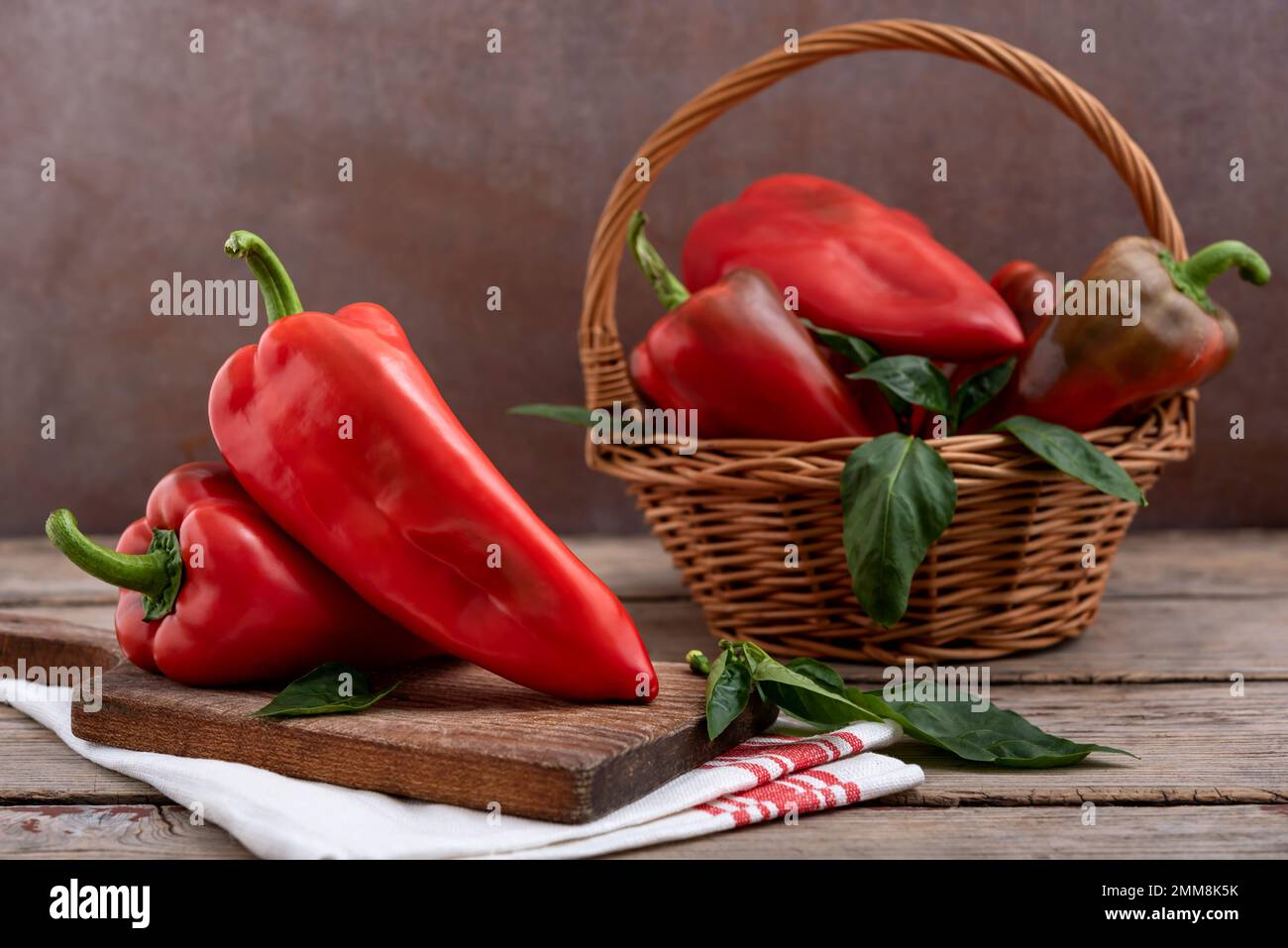 Tagliere e cesto di vimini con peperoni rossi dolci e foglie verdi su tavola di legno. Messa a fuoco selettiva. Foto Stock