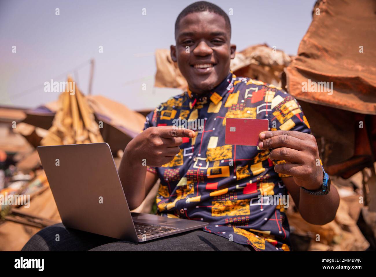 Un ragazzo effettua acquisti online con la sua carta di credito mentre usa il suo computer portatile in un mercato africano Foto Stock