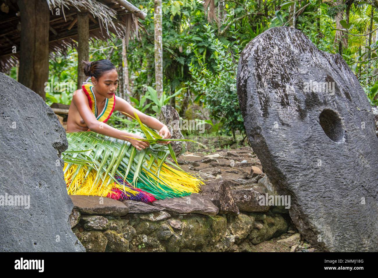 Questa giovane donna (MR) è in un vestito tradizionale per cerimonie culturali e sta tessendo un cesto da una palma frontd e circondato da soldi di pietra in un Foto Stock