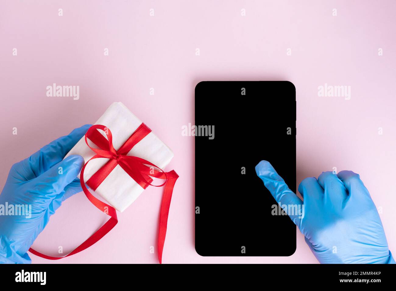 Una mano in un guanto blu protettivo preme su uno schermo nero vuoto di un tablet o telefono, un'altra mano in un guanto protettivo tiene un regalo su una schiena rosa Foto Stock