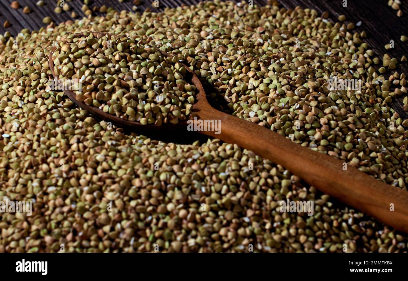 Il grano saraceno verde non lavorato è sparso sul tavolo e un cucchiaio di legno riempito con grani di grano saraceno Foto Stock