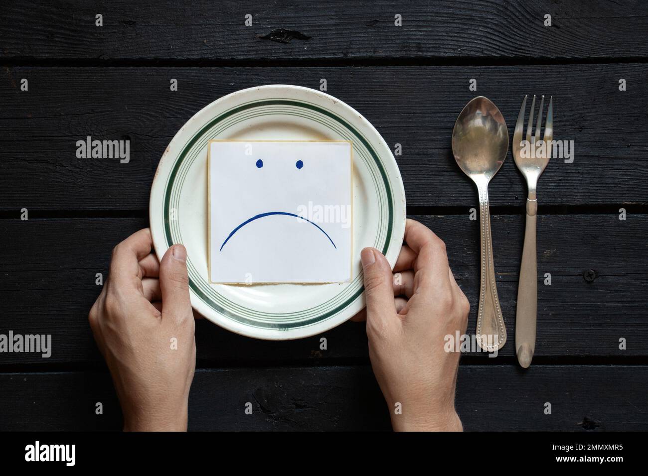 un piatto di carta su cui vengono disegnate una triste faccia sorridente e le mani femminili, fame e niente da mangiare, un cattivo umore a pranzo Foto Stock