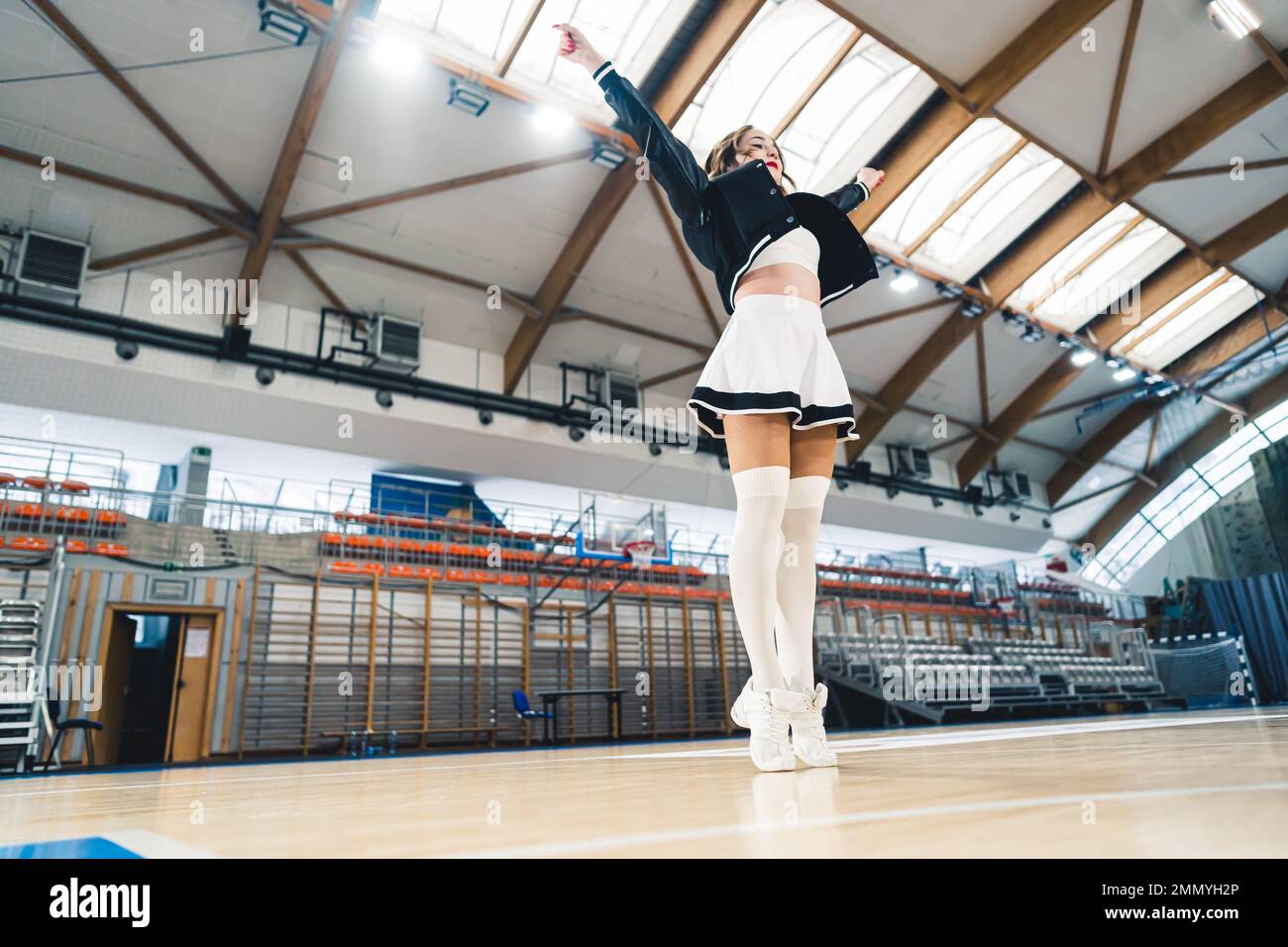 Ragazza cheerleader che fa Y posa in sala sportiva. Campo da pallacanestro sullo sfondo. . Foto di alta qualità Foto Stock