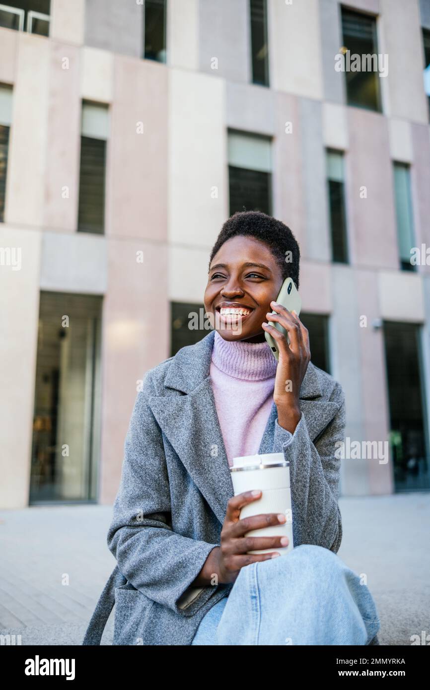 Donna afro-americana felice in outerwear elegante con tazza di caffè eco-friendly. Sta guardando via con il sorriso e sta rispondendo alla chiamata telefonica sulla strada della città di mattina Foto Stock