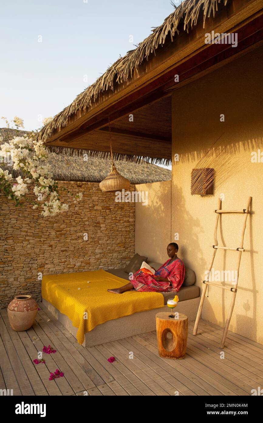 Senegal, il delta del Saloum dichiarato Patrimonio Mondiale dall'UNESCO, Palmarin, Yokan Lodge, donna in un abito colorato con un libro in mano sdraiato su un letto sulla terrazza della sua suite in tonalità ocra Foto Stock