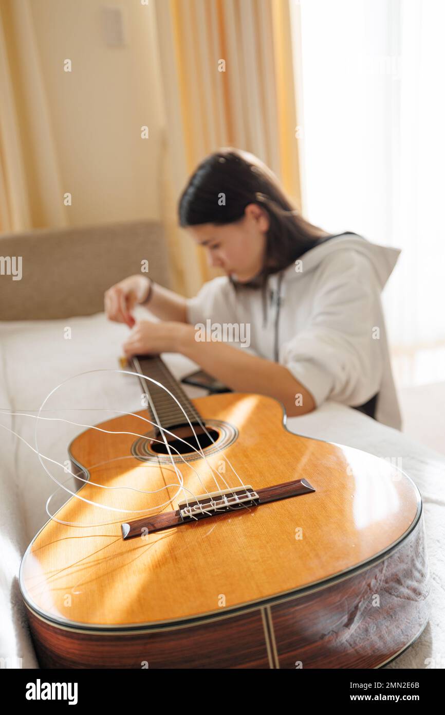 Ragazza adolescente con lunghi capelli castani e zoccoli bianchi che cambiano le corde in chitarra acustica classica mentre si siede sul pavimento. Foto Stock