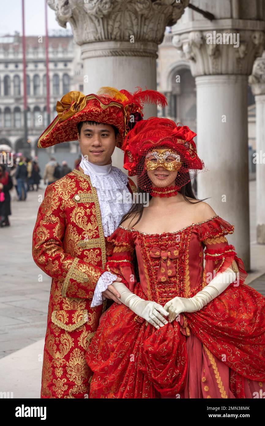 Giovane asiatico e giovane donna in costume di carnevale rosso medievale, cappelli con piume e maschere in Piazza San Marco a Venezia Foto Stock