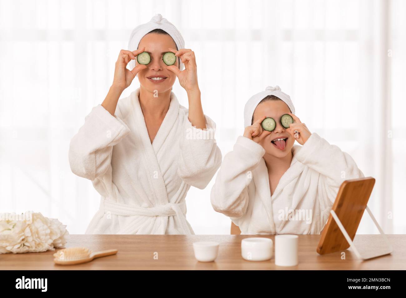 Allegra mamma e bambina indossando accappatoi che applicano fette di cetriolo sugli occhi Foto Stock