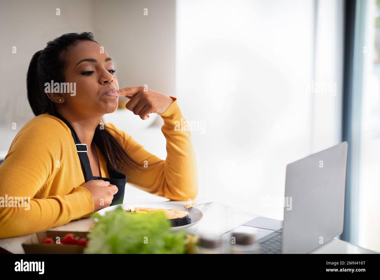 Felice millenario afroamericana donna in grembiule cercando piatto di pesce, leccando dito, guardando video blog sul computer portatile Foto Stock