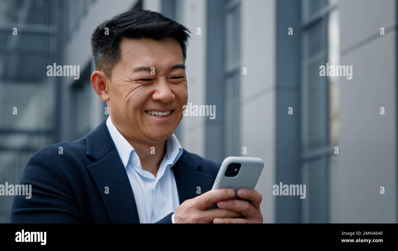Emotivo sorridente ridere asiatico di mezza età uomo d'affari coreano imprenditore imprenditore datore di lavoro risata guardando il cellulare chattando navigazione sorriso Foto Stock