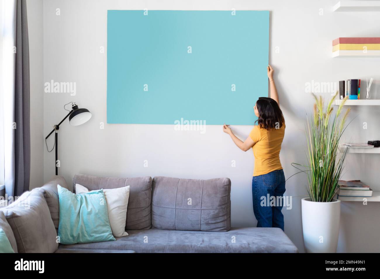 Giovane donna che appende un grande poster vuoto sulla parete nel soggiorno Foto Stock
