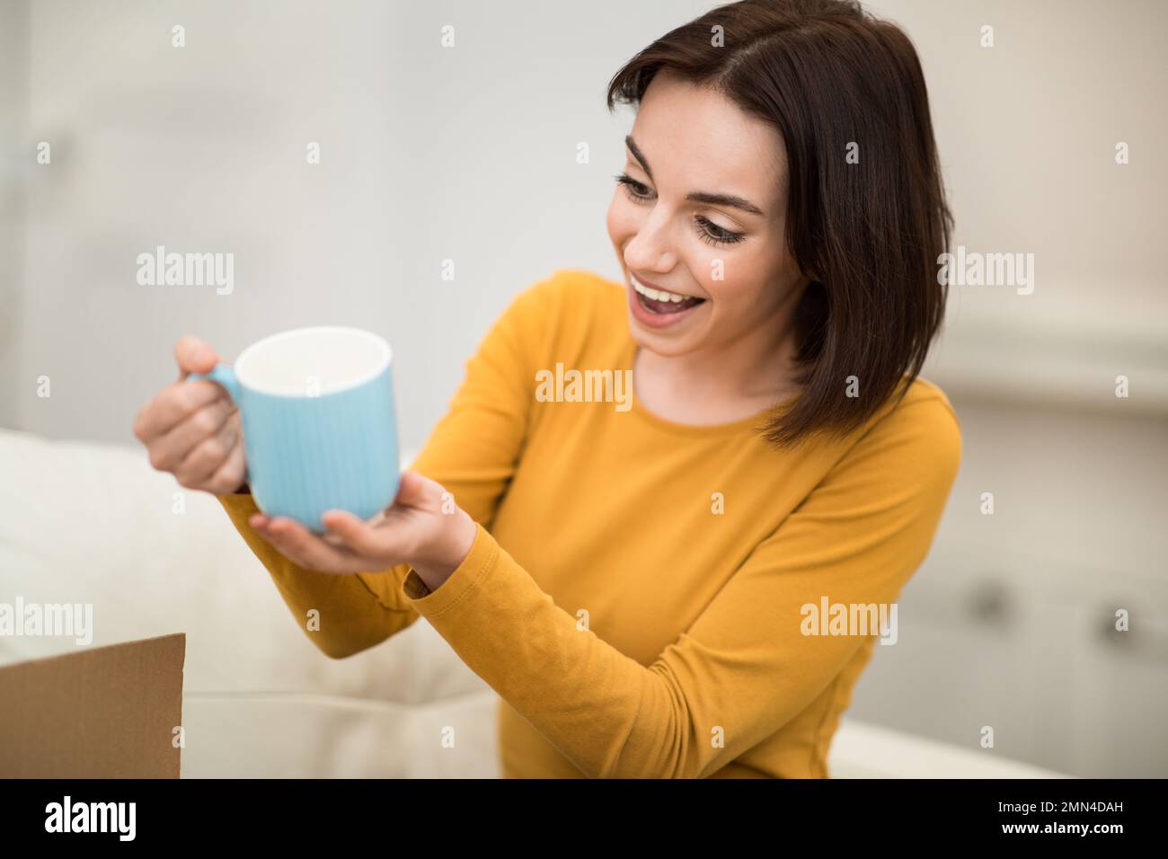 Giovane donna eccitata che tiene tazza blu, interno della casa Foto Stock