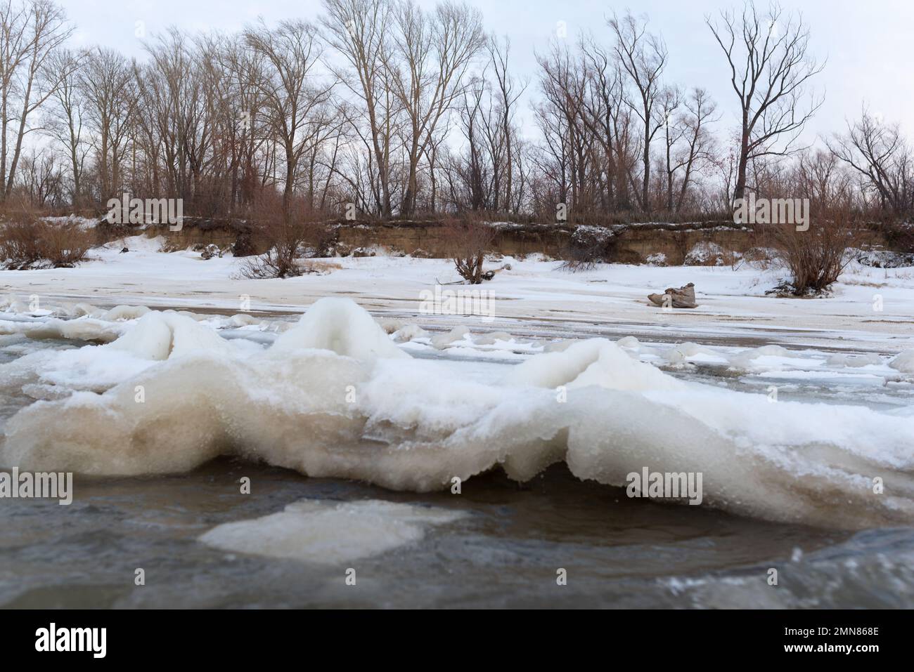 Il ghiaccio e la neve sulla riva del fiume si innalza in onde dal vento e le correnti d'acqua accanto agli alberi sulla scogliera della riva. Foto Stock