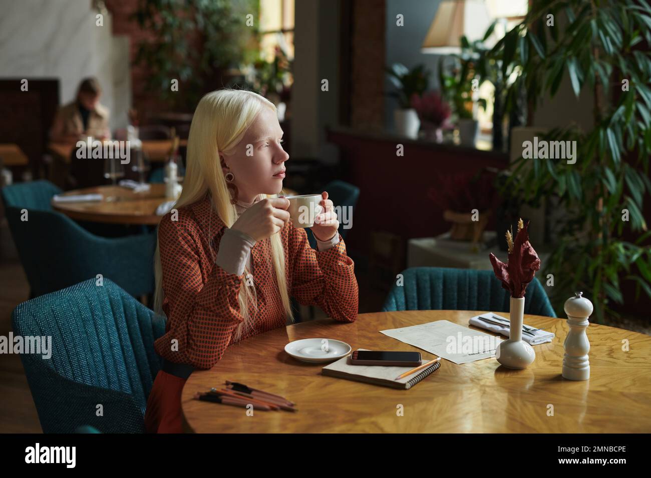 Giovane donna albina pensiva con tazza di tè seduto in comoda poltrona da tavolo in accogliente caffè decorato con piante verdi Foto Stock