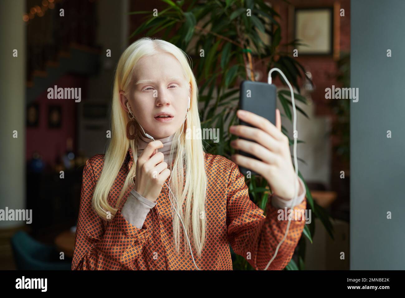 Giovane ragazza albino in auricolari che tiene il telefono cellulare di fronte a se stessa mentre guarda il suo amico sullo schermo durante la comunicazione in video chat Foto Stock