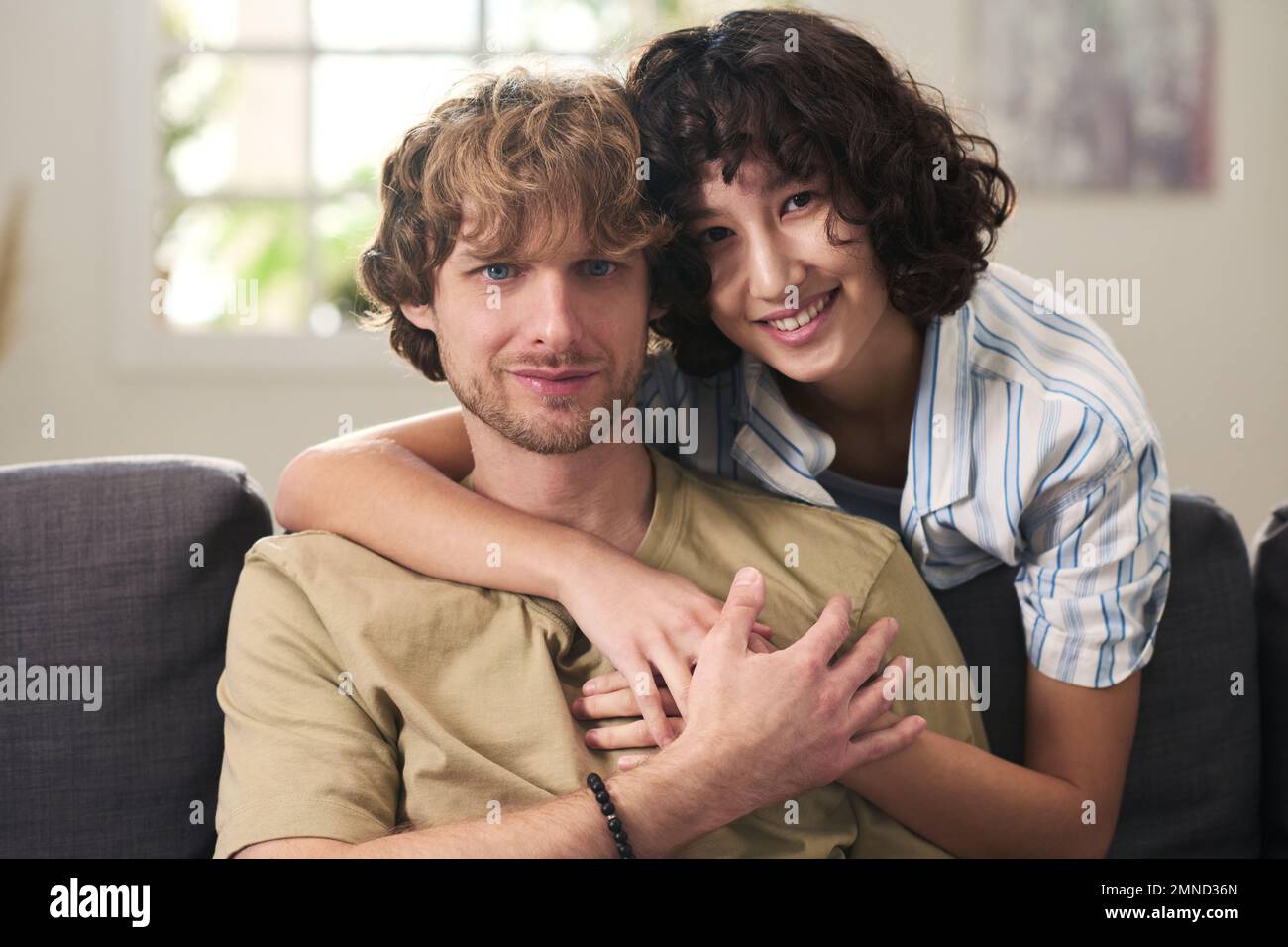 Giovane coppia affettuosa che si rilassa sul divano e guarda la macchina fotografica mentre felice bruna donna che si piega sopra il marito e lo abbraccia Foto Stock