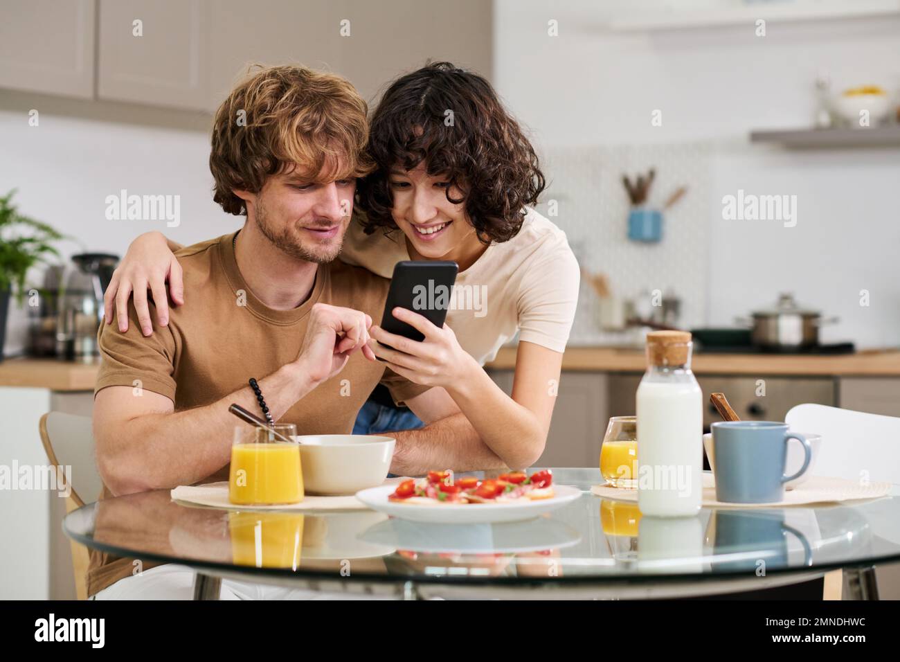 Giovane donna sorridente che mostra qualcosa in smartphone a suo marito seduto al tavolo con panini, succo di frutta e latte e fare colazione Foto Stock