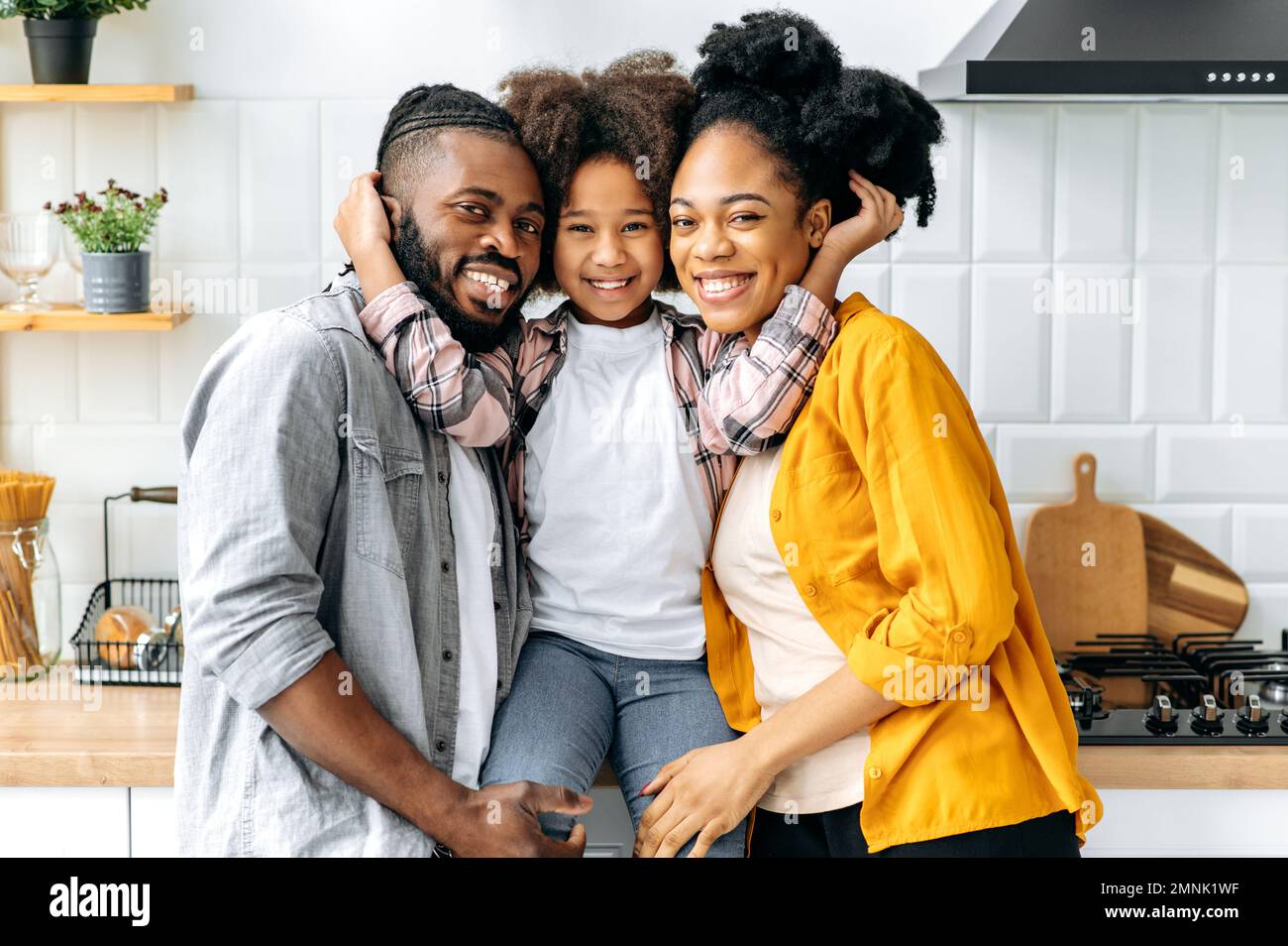 Ritratto di famiglia. African American felice famiglia, papà mamma e la loro cute prescolare figlia, abbracciando insieme in una cucina a casa, guardare la macchina fotografica, sorridere, felice di essere insieme, ragazza abbracciando i suoi genitori Foto Stock