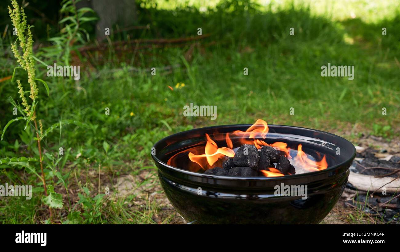 Barbecue nero rotondo con fuoco all'aperto con erba verde. Fuoco fiamma. Carbone di riparazione per grigliare il barbecue all'aperto Foto Stock