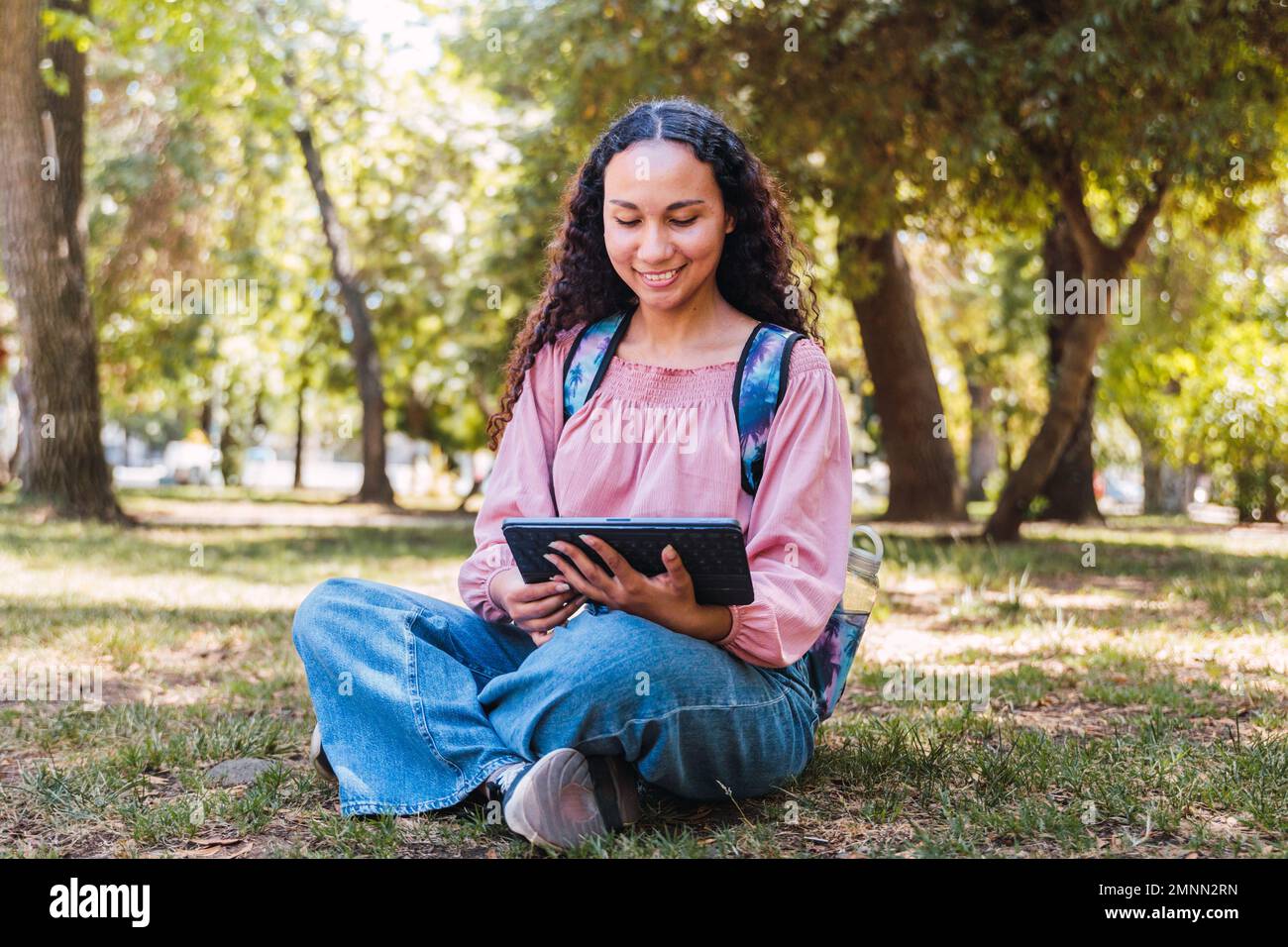 Studentessa universitaria latina sorridente e con un tablet seduto all'aperto in un parco sull'erba Foto Stock