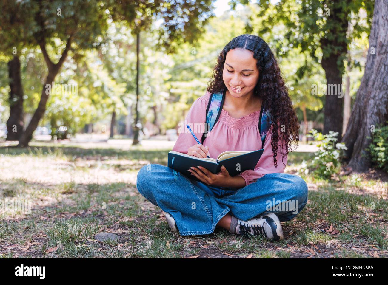 Studentessa universitaria latina che sorride e scrive a mano nel suo diario, sull'erba in un parco Foto Stock