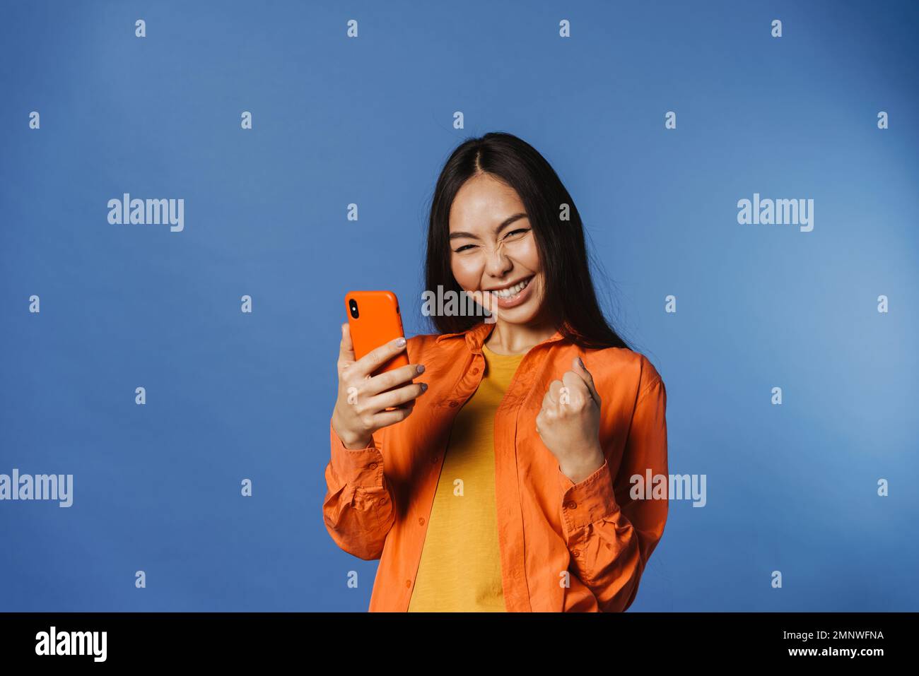 Giovane donna asiatica che indossa una camicia che stringe il pugno mentre usa il cellulare isolato su sfondo blu Foto Stock