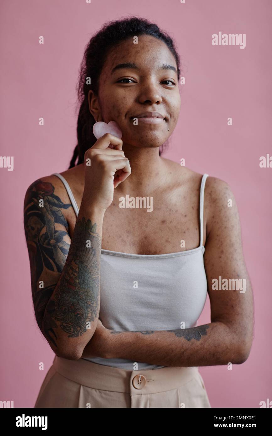 Ritratto verticale di giovane donna nera con cicatrici acne utilizzando il massaggiatore facciale e sorridendo contro il rosa Foto Stock