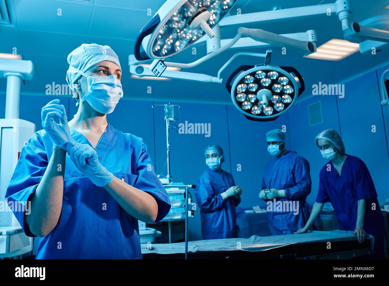 Infermiere della sala operatoria che indossa guanti protettivi prima dell'intervento chirurgico in piedi in una sala operatoria moderna con personale medico Foto Stock