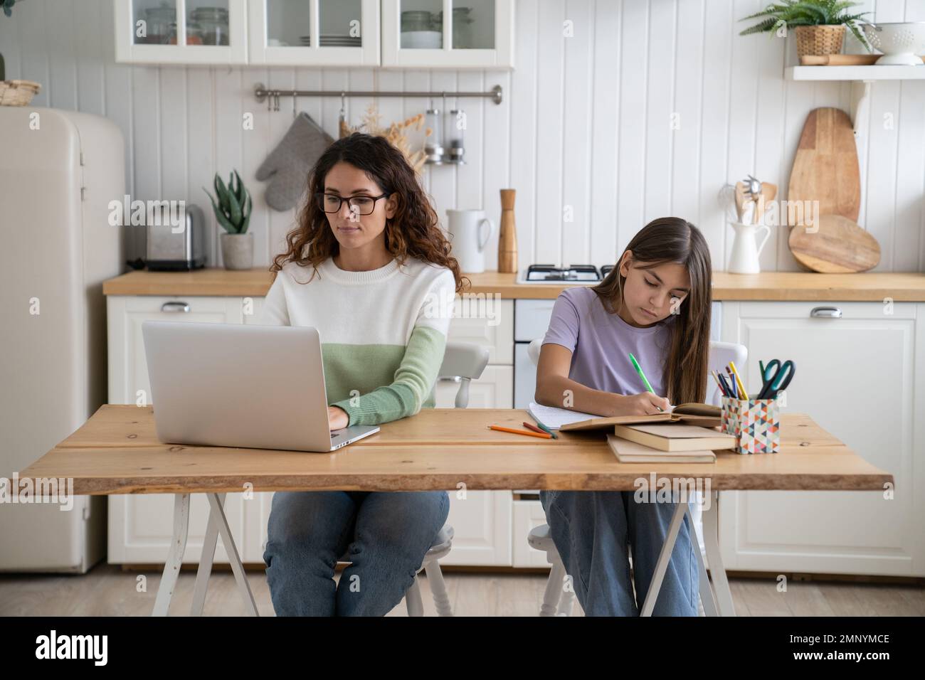 Donna concentrata madre siede al tavolo da cucina lavoro a distanza sul portatile vicino con ragazza adolescente Foto Stock