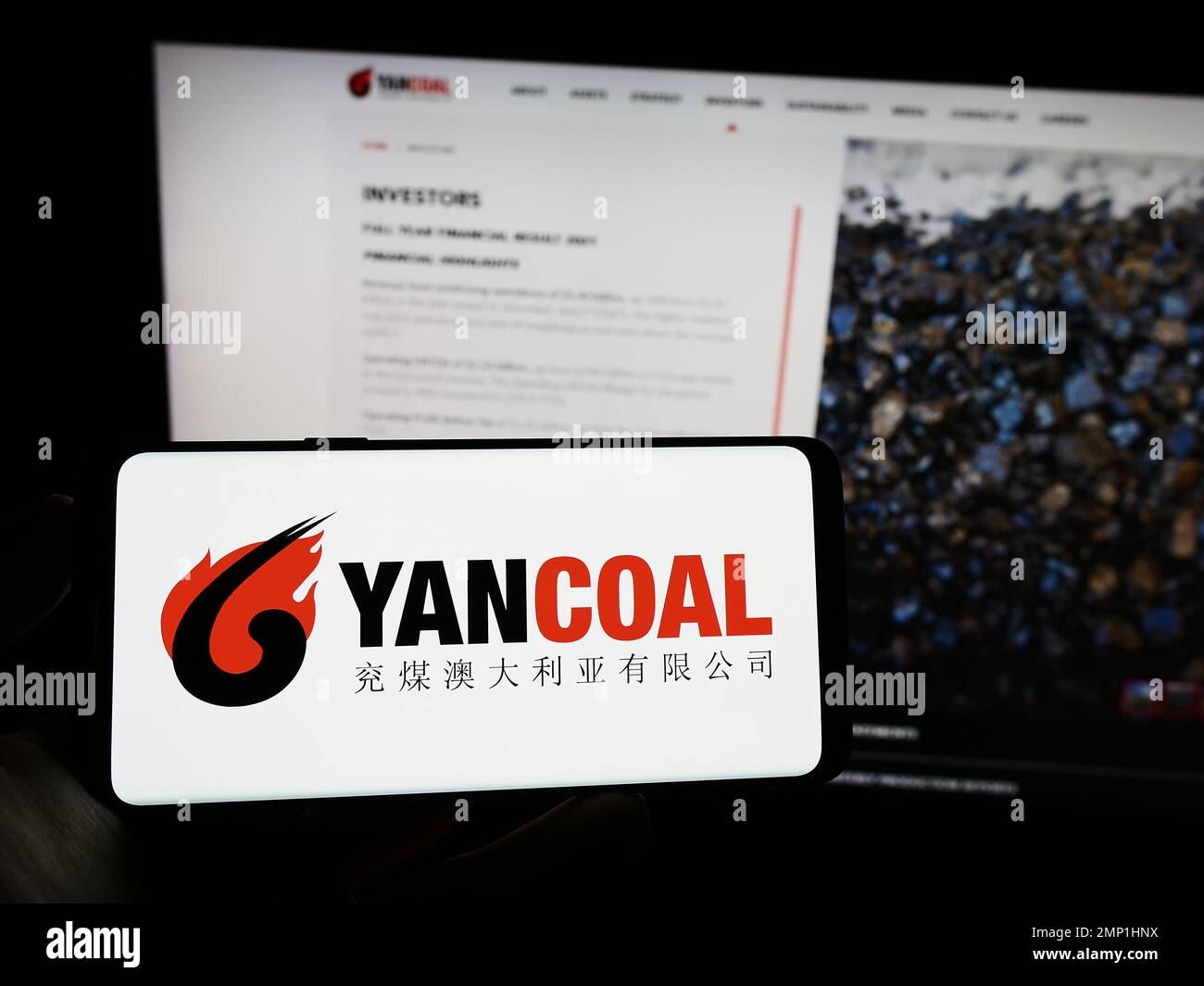 Persona che tiene il cellulare con il logo della società di estrazione del carbone Yancoal Australia Limited sullo schermo di fronte al logo aziendale. Messa a fuoco sul display del telefono. Foto Stock
