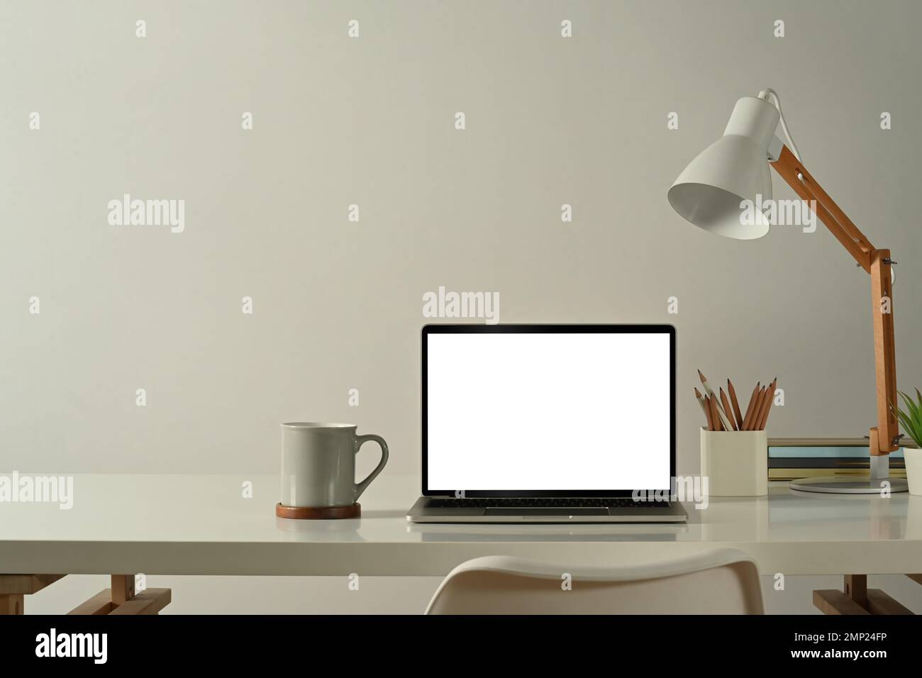 Vista frontale del computer portatile, della tazza da caffè, del portamatite e della lampada sul tavolo bianco in un ufficio domestico di dimensioni ridotte Foto Stock