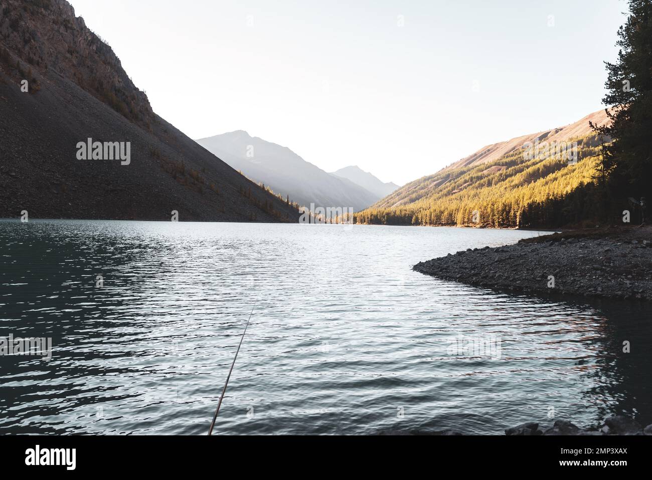 Una canna da pesca si trova all'ombra delle rocce sulla riva del lago alpino Shavlinskoe la sera con la luce del sole sulle montagne all'orizzonte. Foto Stock