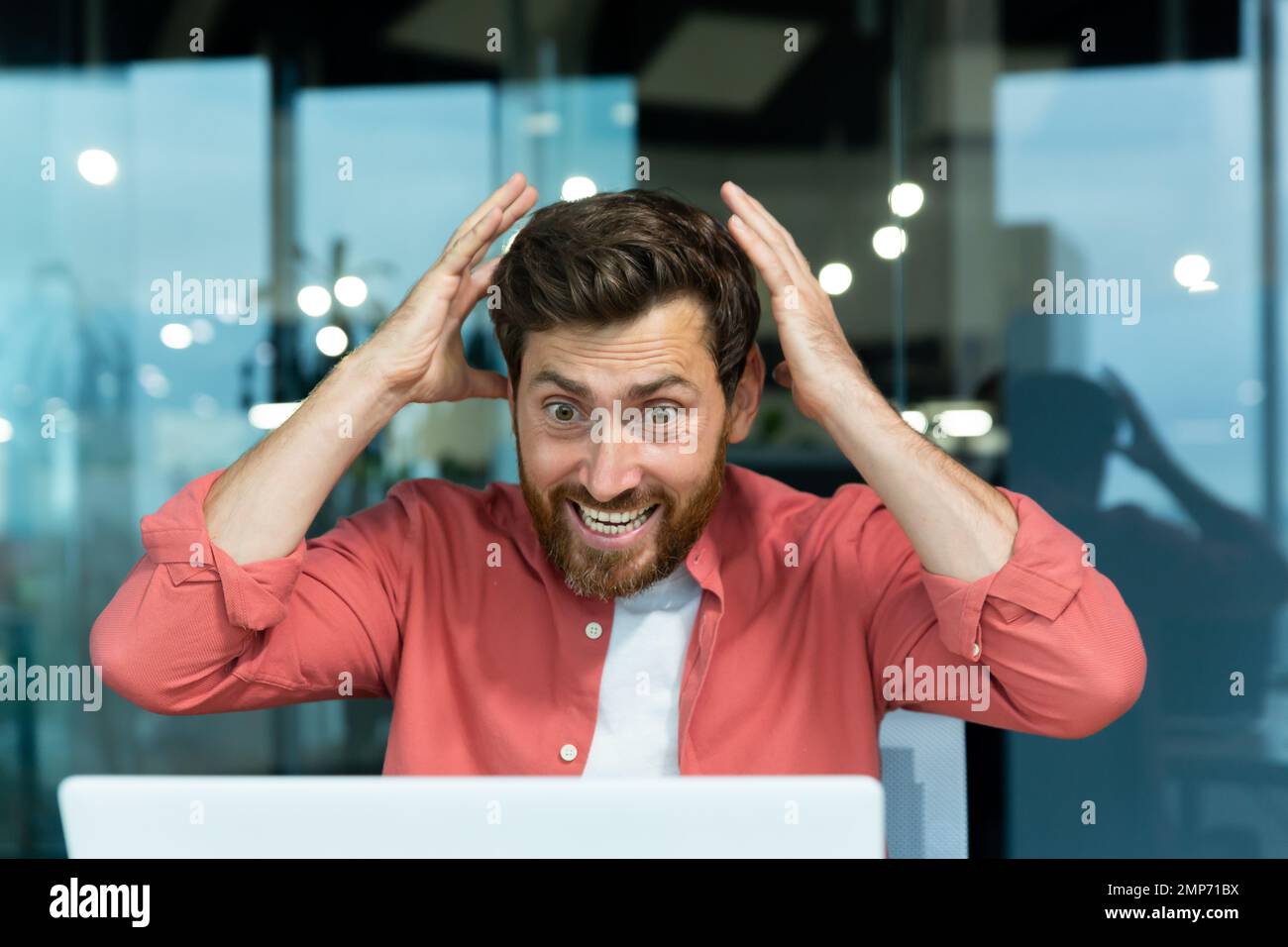 Primo piano. Uomo scioccato e felice in camicia rossa seduto alla scrivania in ufficio e guardando il portatile. Grida di gioia, afferra la testa con le mani. Foto Stock