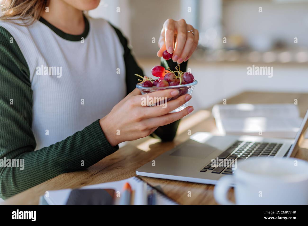 Vista laterale della donna che tiene il recipiente con uva sopra la scrivania con computer, diario e smartphone. Concetto di equilibrio tra vita lavorativa e vita privata. Foto Stock