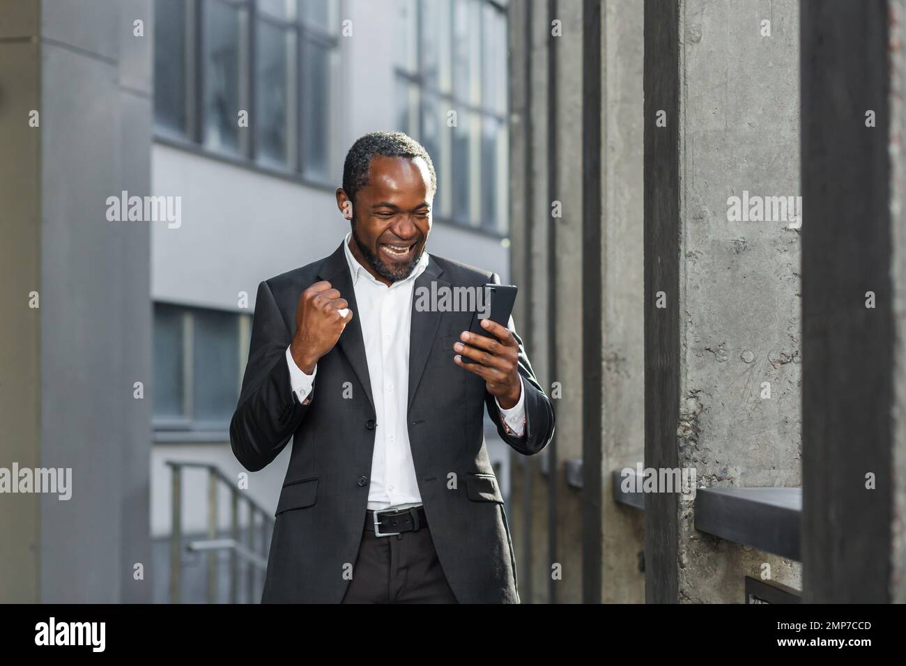 Felice giovane uomo afroamericano in piedi vicino al centro ufficio in tuta. Tiene il telefono nelle sue mani, guarda il display, festeggia, esulta. Ho ricevuto buone notizie, messaggi, vincite. Foto Stock