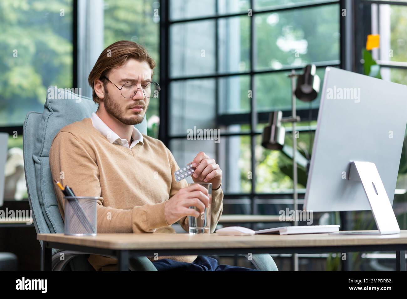 Lavoratore di ufficio malato e troppo stanco al lavoro con un computer, uomo d'affari all'interno dell'ufficio prende una pillola medica e bevande acqua, un uomo biondo in abiti casual, un maglione con una camicia. Foto Stock