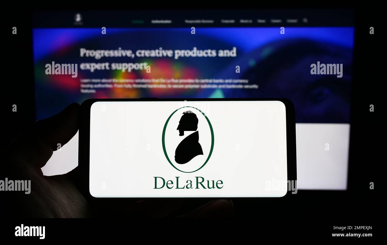 Persona che tiene uno smartphone con il logo del produttore di banconote britanniche De la Rue plc sullo schermo di fronte al sito Web. Messa a fuoco sul display del telefono. Foto Stock