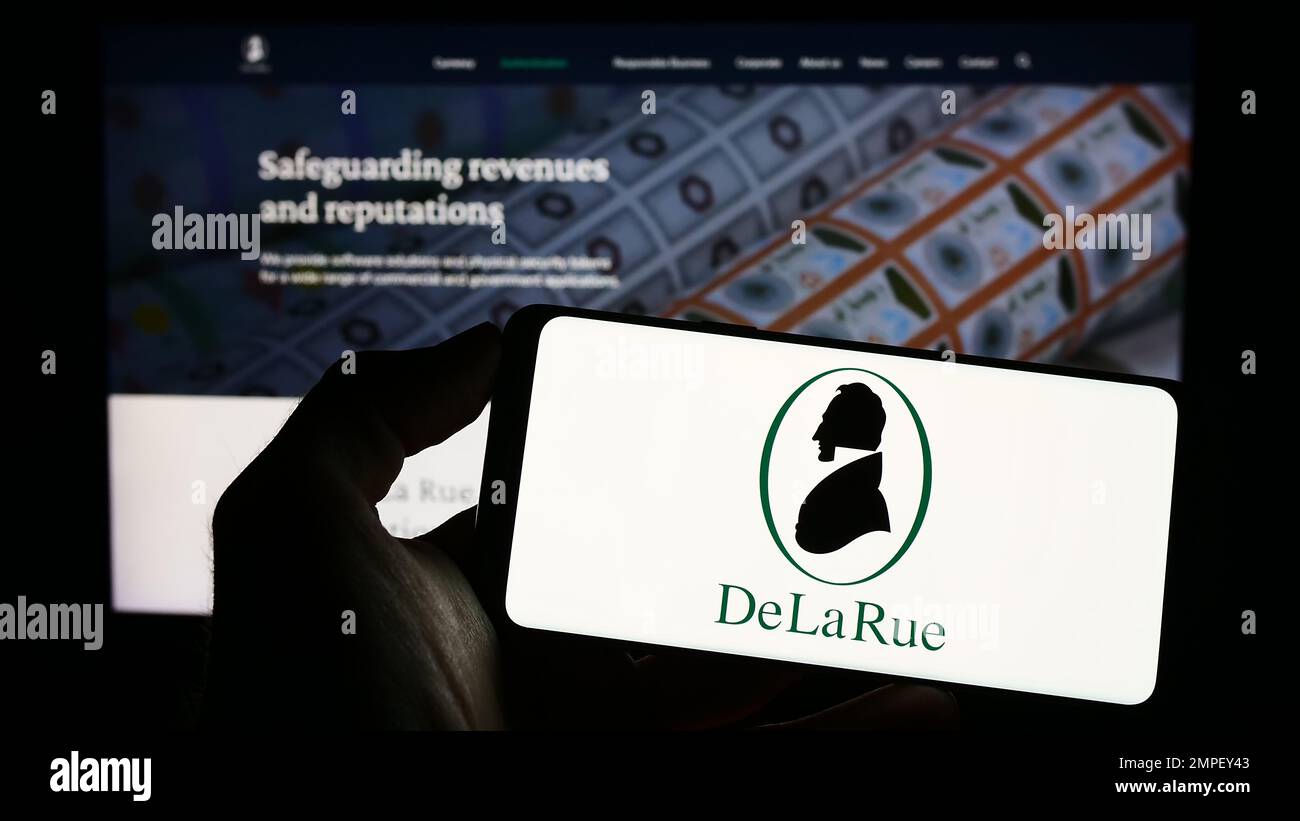 Persona che tiene il cellulare con il logo del produttore britannico di banconote De la Rue plc sullo schermo di fronte alla pagina web aziendale. Messa a fuoco sul display del telefono. Foto Stock