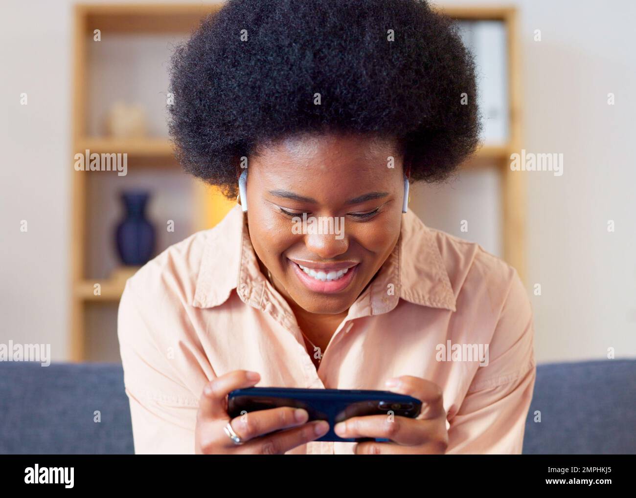 Donna che gioca un gioco per cellulare sul suo telefono in modalità orizzontale nel salotto di casa. Giovane giocatore eccitato sorridente e ridendo mentre godendo un certo divertimento Foto Stock