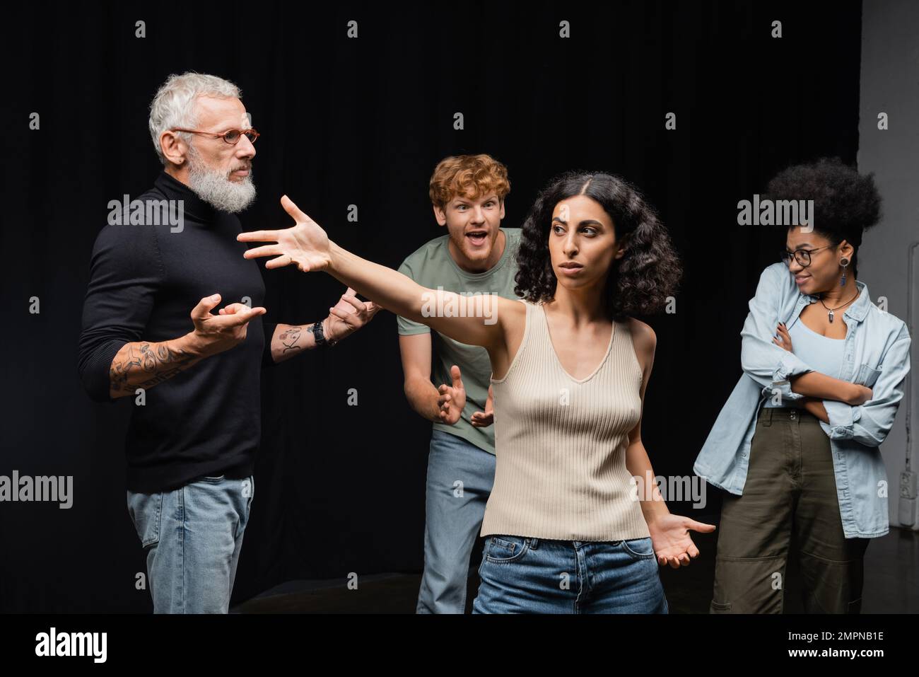 donna multirazziale in piedi con mano tesa vicino al direttore d'arte barbuto e gli studenti interrazziali emotivi nella scuola di recitazione Foto Stock