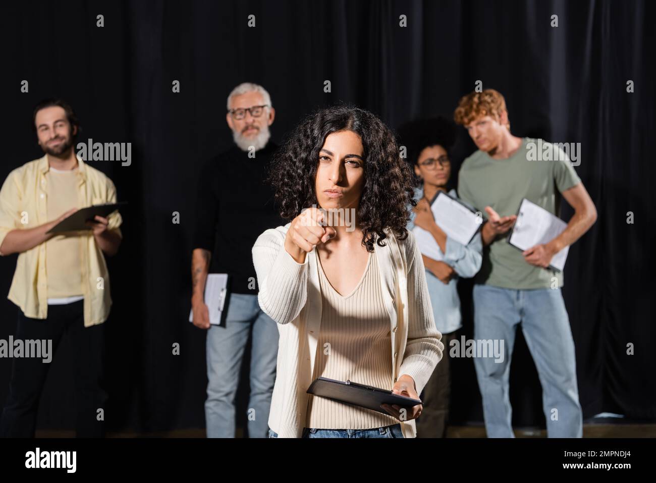 donna multirazziale con un'espressione del volto seria che indica la telecamera mentre si prova vicino ad attori sfocati e a un direttore artistico maturo Foto Stock