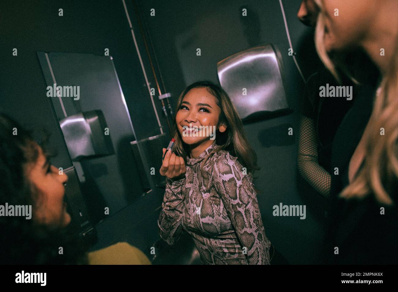 Giovane donna sorridente che applica il rossetto mentre guarda gli amici nella stanza da bagno del nightclub Foto Stock