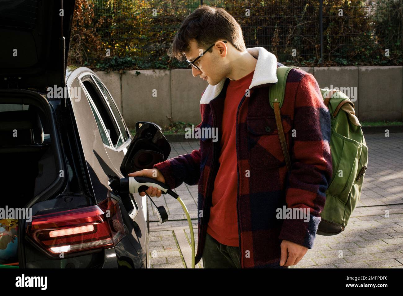 Uomo che indossa lo zaino mentre carica l'auto elettrica alla stazione Foto Stock