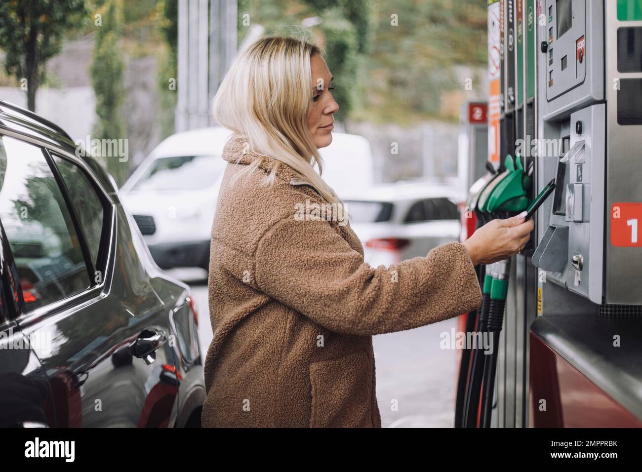 Vista laterale della donna bionda che effettua il pagamento tramite smartphone mentre si trova in piedi presso la stazione di rifornimento Foto Stock