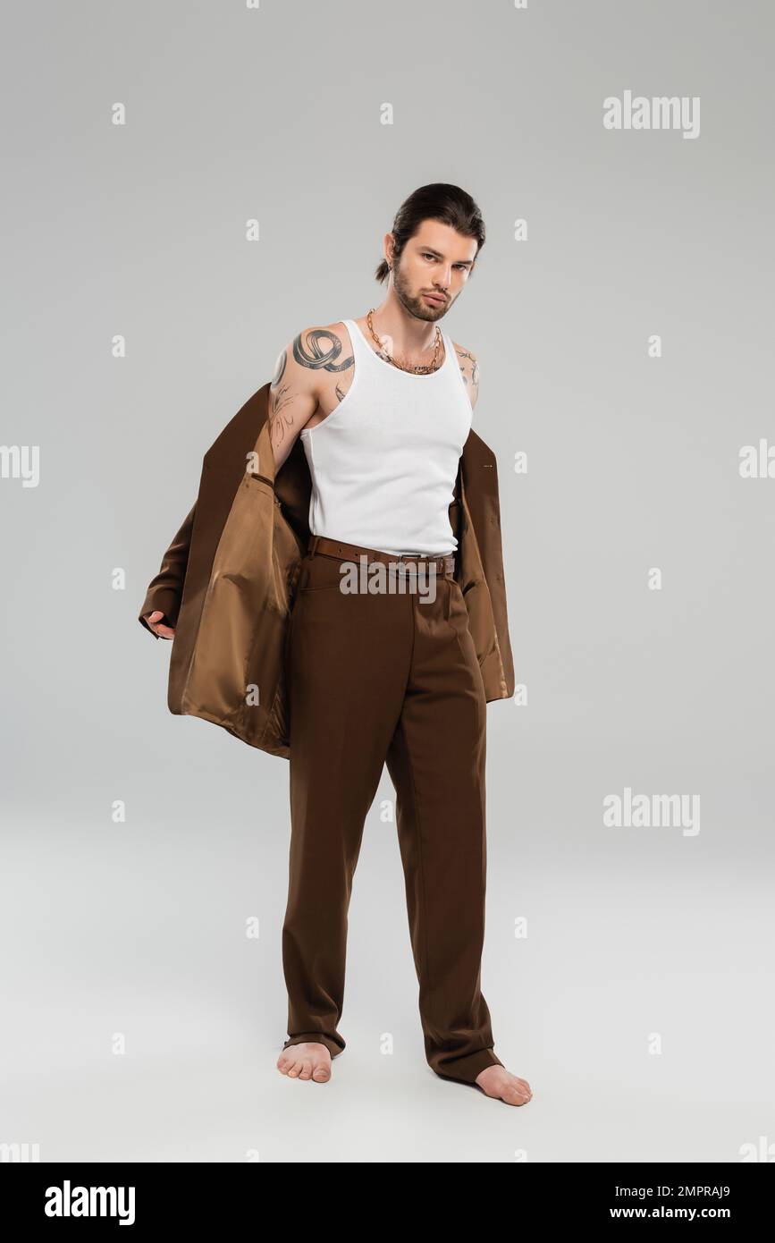 Tutta la lunghezza dell'uomo tatuato a piedi nudi che togli la giacca su sfondo grigio Foto Stock