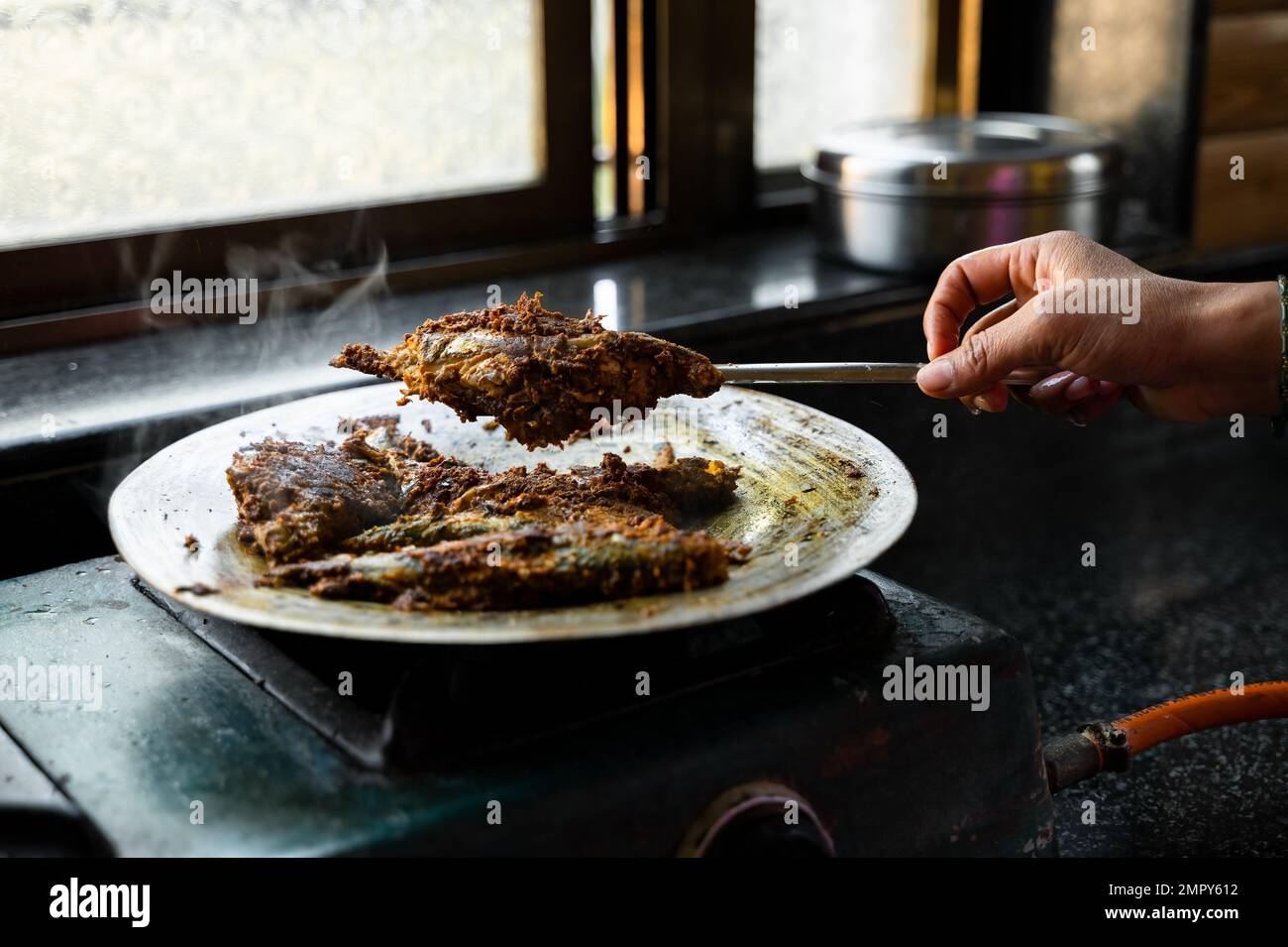 Sgombro indiano Pesce marinato con spezie friggere su metallo su alluminio utensile e un pezzo di pesce sollevato con l'aiuto di cucchiaio con mano donna. Foto Stock