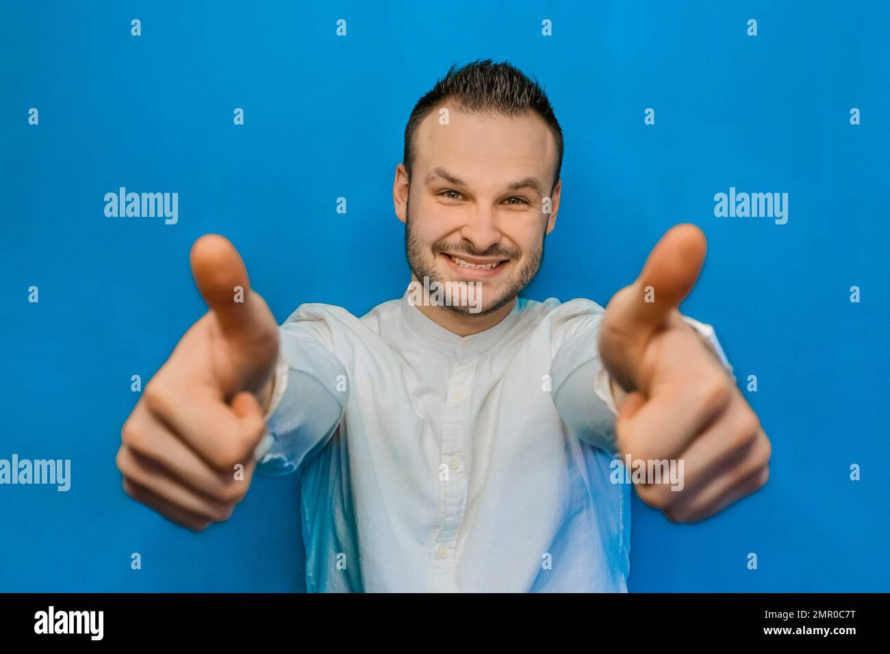 Ritratto di giovane attraente uomo d'affari felice europeo in camicia bianca con barba che mostra i pollici cool su due mani su sfondo blu. Foto Stock
