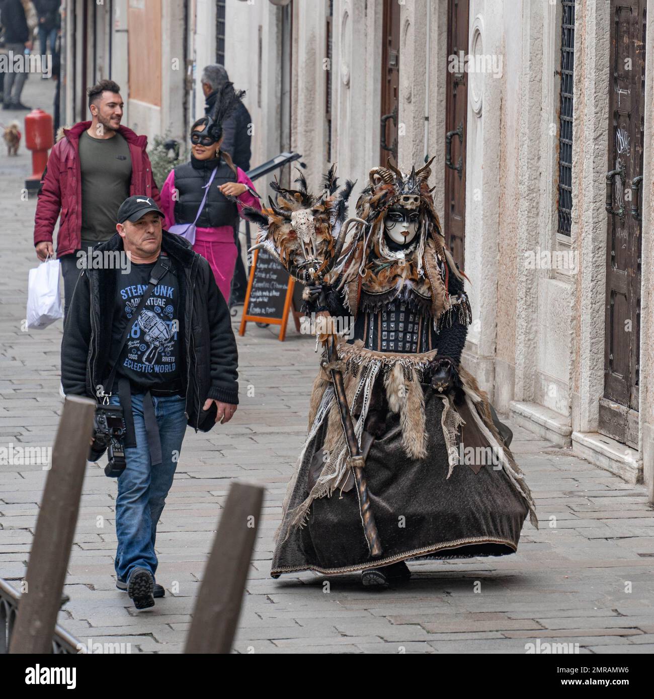 Il fotografo cammina per strada con un modello vestito in costume carnevale di strega pagana con le sculture, le pellicce e le piume su di essa a Venezia Foto Stock