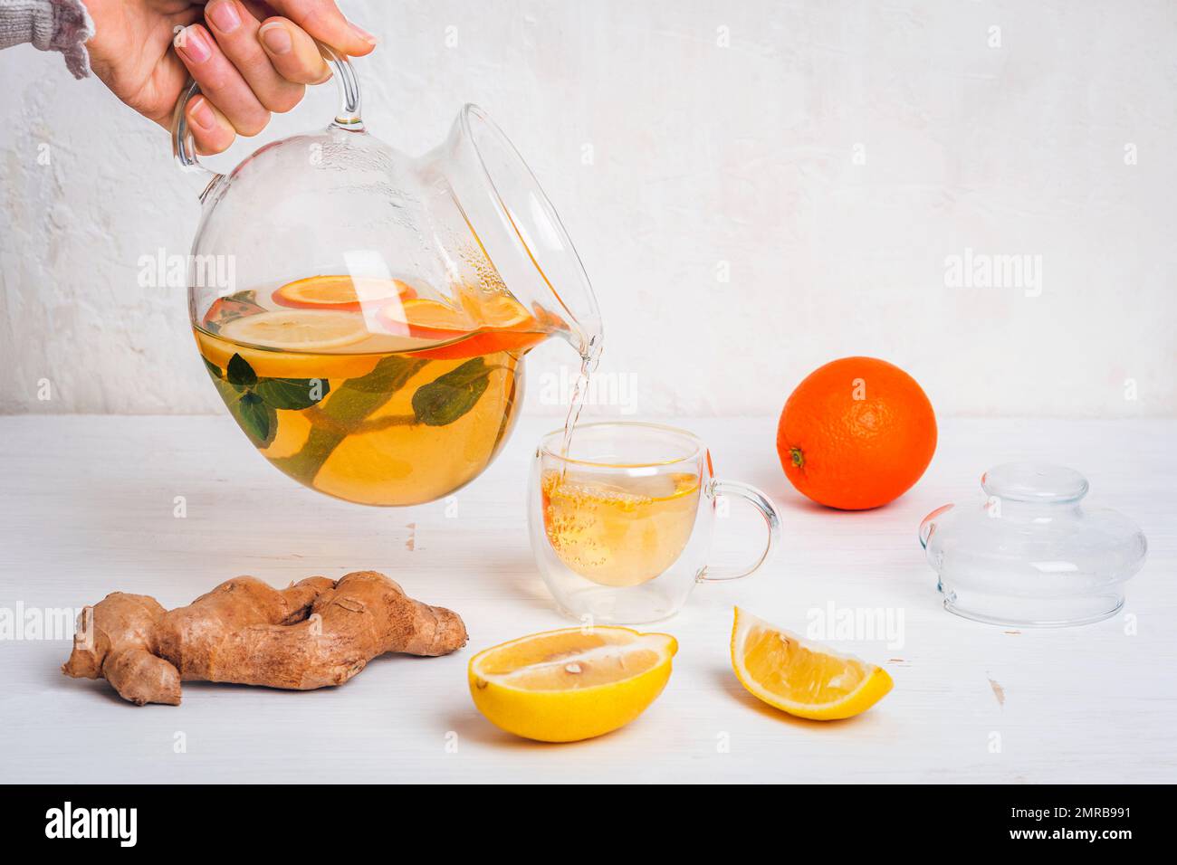 Teiera di vetro con tè all'arancia, al limone e allo zenzero che si versa in una tazza. Ingredienti su tavola di legno bianco. Foto Stock