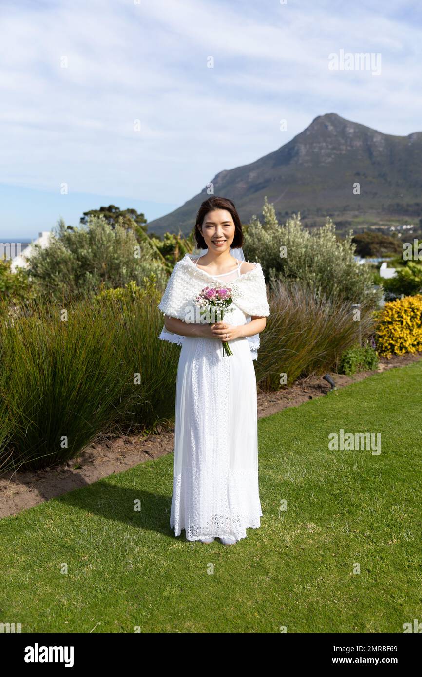Ritratto verticale di felice sposa asiatica che tiene bouquet sorridente al matrimonio all'aperto, spazio copia Foto Stock Ritratto verticale di felice sposa asiatica che tiene bouquet sorridente al matrimonio all'aperto, spazio copia Foto Stock